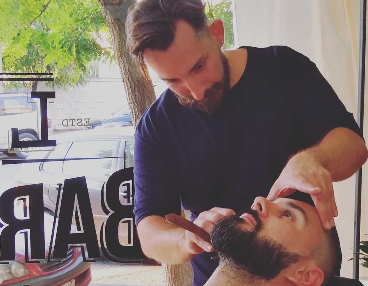 Ο barber του LIORAS BARBERSHOP περιποιείται τη γενειάδα πελάτη με ξυράφι, κρατώντας του το κεφάλι με το άλλο χέρι, μπροστά στην τζαμαρία του μαγαζιού με το φως του δρόμου