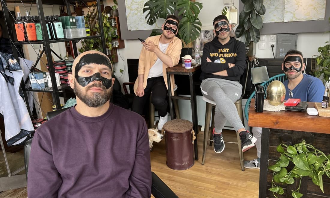 Τέσσερις πελάτες του LIORAS BARBERSHOP με μαύρη μάσκα άνθρακα στο πρόσωπο, καθισμένοι στον χώρο του μαγαζιού ανάμεσα σε φυτά, ένας με το κινητό του και ένας σκύλος στο πάτωμα